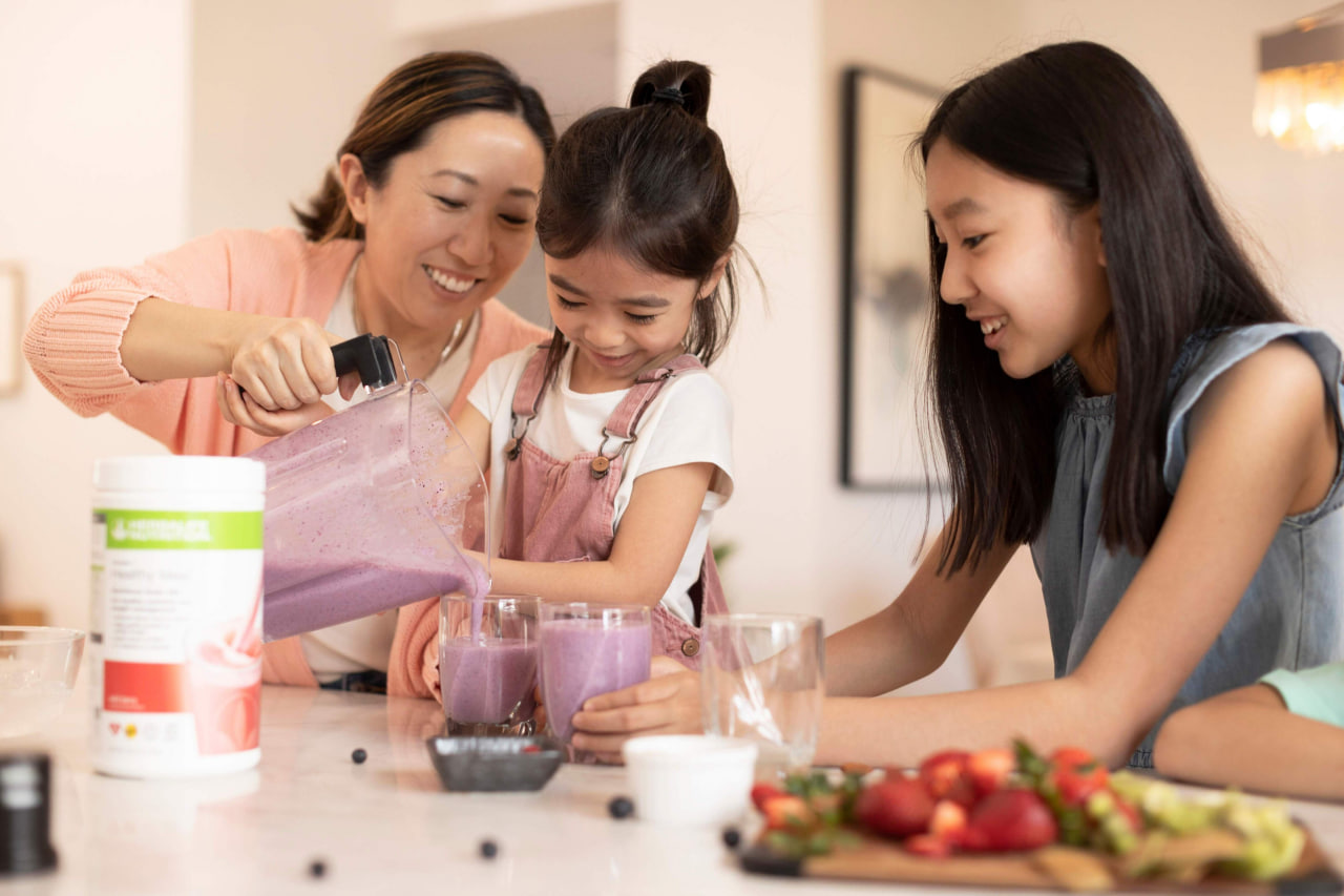 Seorang wanita bersama dua kanak-kanak menuang minuman smoothie ungu ke dalam gelas di sebelah produk Herbalife